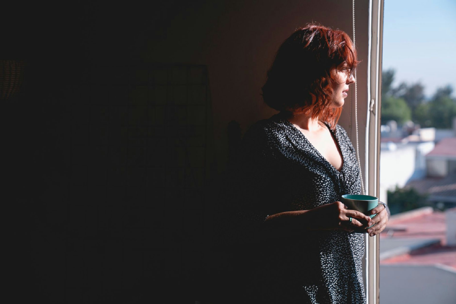 Mulher praticando a autocompaixão, olhando pela janela de forma serena em sua casa em Curitiba, refletindo sobre seu bem-est…