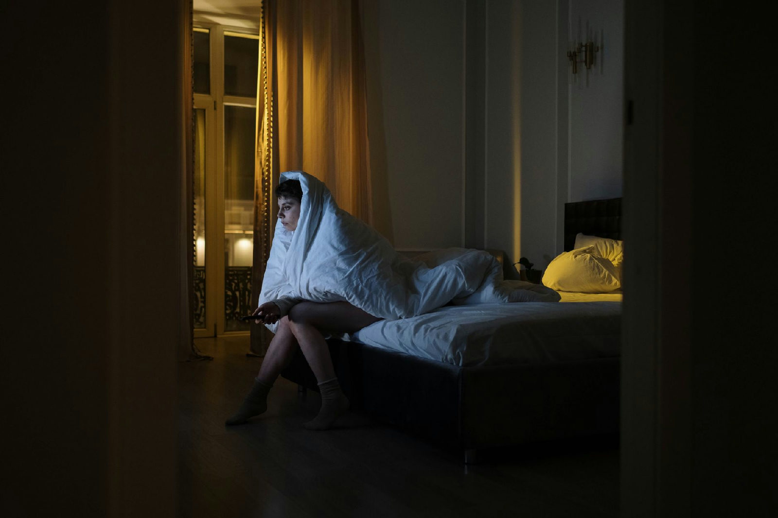 Mulher sentada na cama à noite, com expressão calma, olhando pela janela, ilustrando como acalmar os pensamentos acelerados…